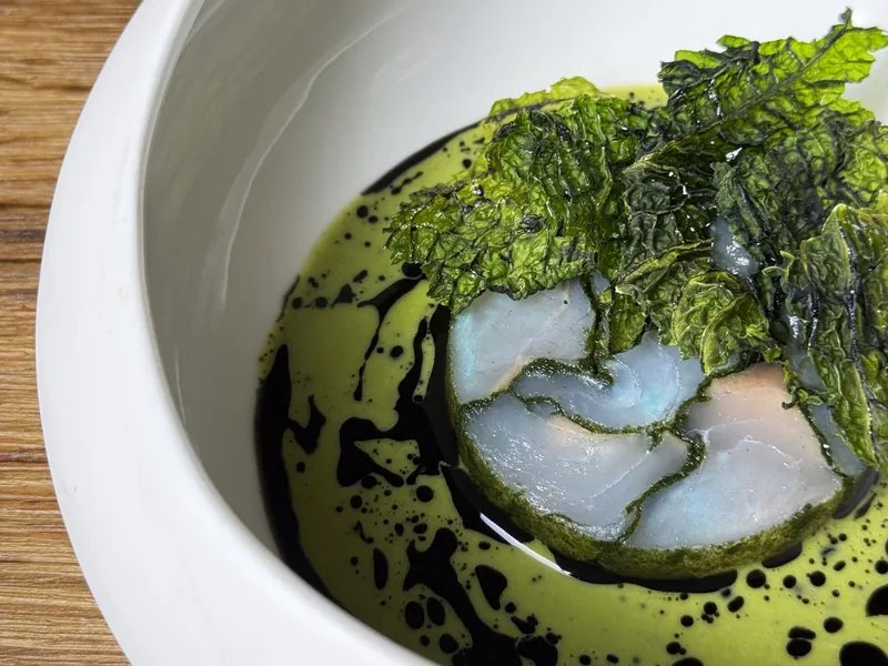 Een gerecht in een witte kom: groene bladeren in een groene saus met donkere druppels